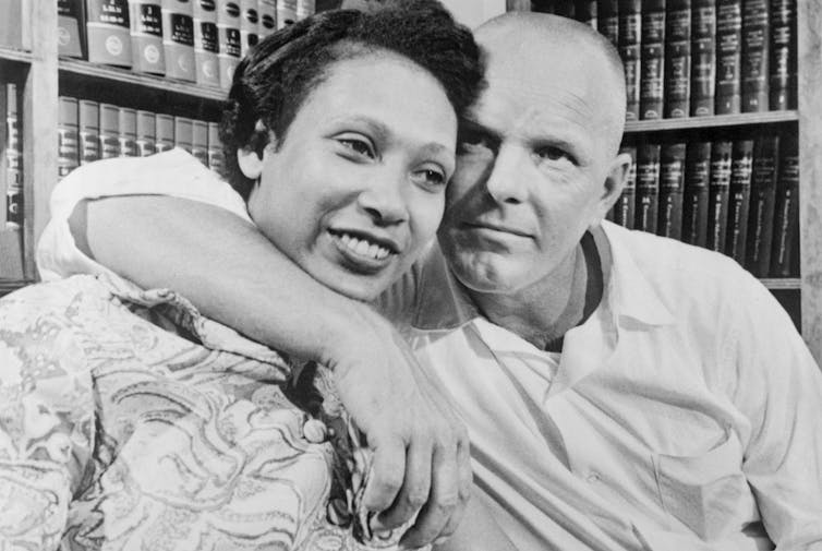 A black and white photo shows a woman sitting next to a man, with his arm around her and their heads touching.