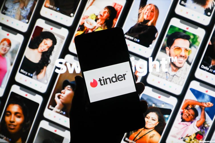 A phone is black except for a white rectangle and the word “Tinder” and lies in front of many other phone screens with photos of various people.