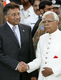 Man in dress suit wearing glasses shakes hands with man wearing white suit.
