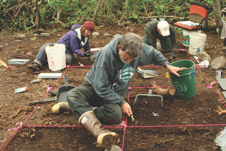 three people seated on ground use hand tools to dig in the brown earth
