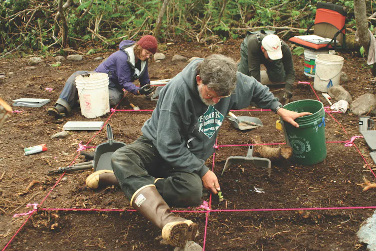 three people seated on ground use hand tools to dig in the brown earth