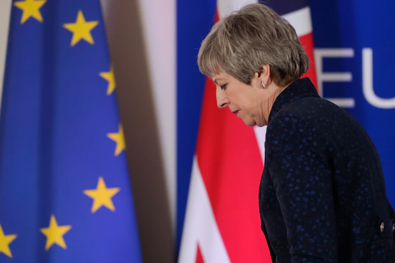 Theresa May sadly walking in front of an EU and UK flag