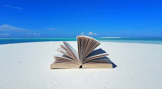 An open book lies on a white sandy beach against the backdrop of a mostly clear blue sky.