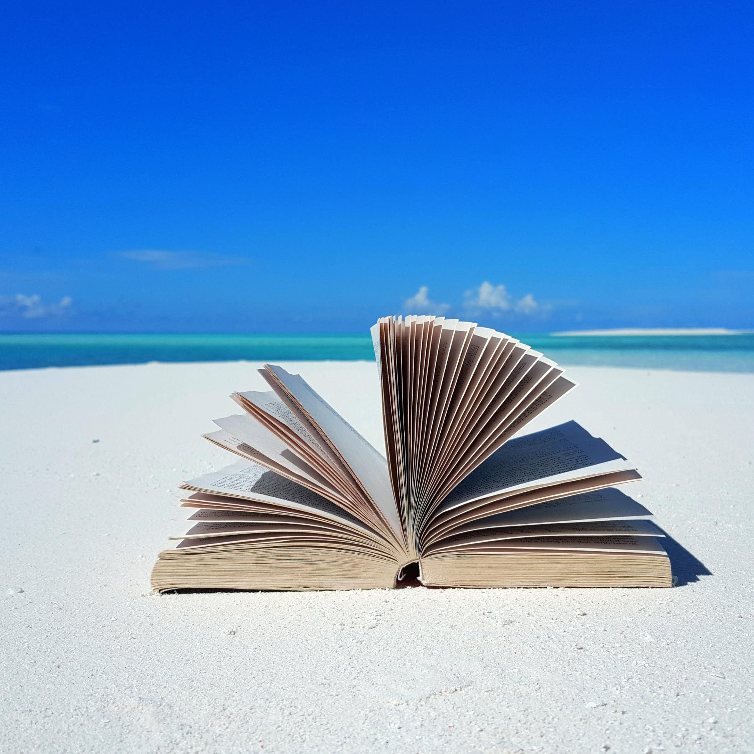 An open book lies on a white sandy beach against the backdrop of a mostly clear blue sky.