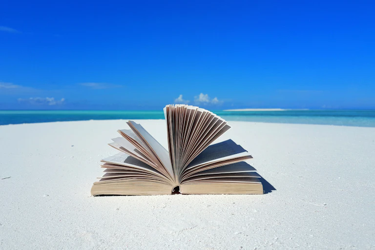 An open book lies on a white sandy beach against the backdrop of a mostly clear blue sky.