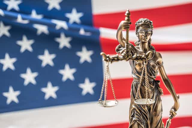 A statue of the goddess of justice, blindfolded and holding scales and a sword, in front of a U.S. flag