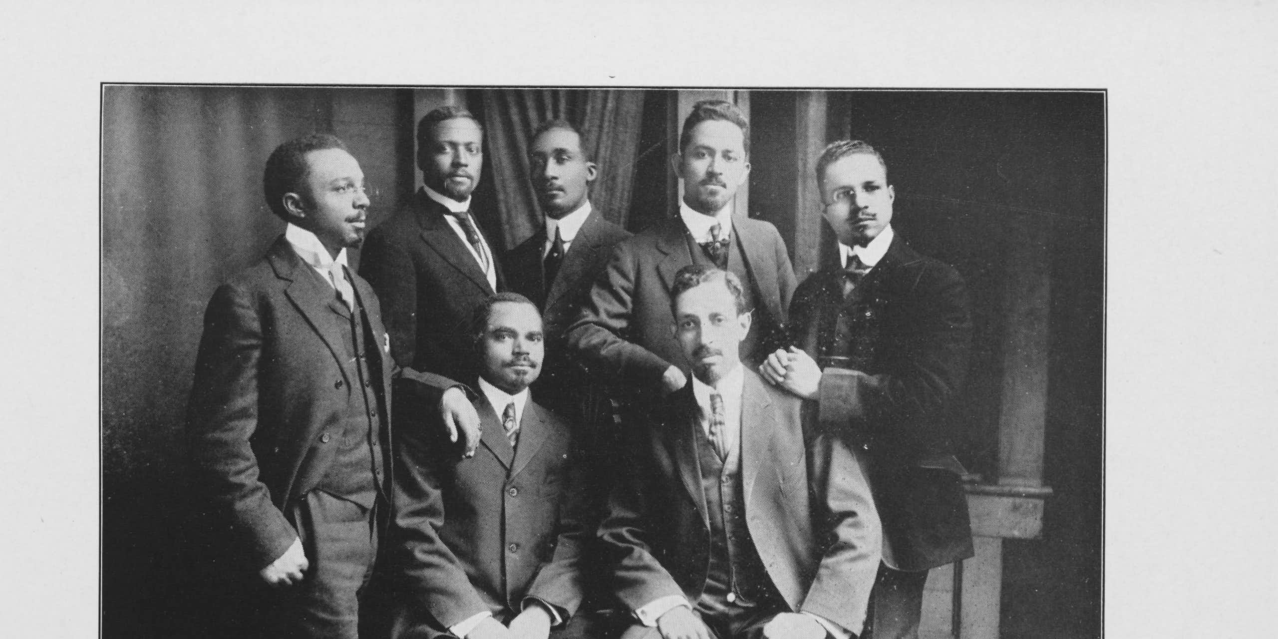 A group of seven Black men are dressed in business suits.