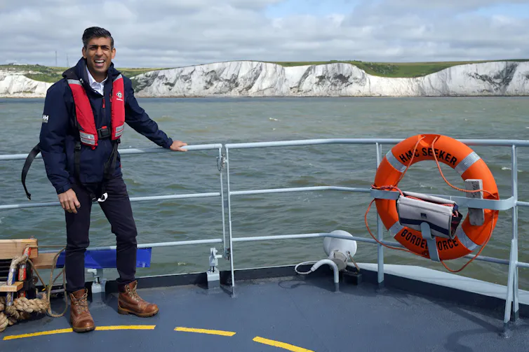Rishi Sunak stands smiling on the deck of a boat with a view of the Cliffs of Dover behind him.