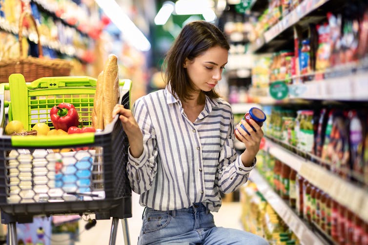 A young woman crouched down beside a full shopping cart, looking at the label on a can in a grocery store aisle