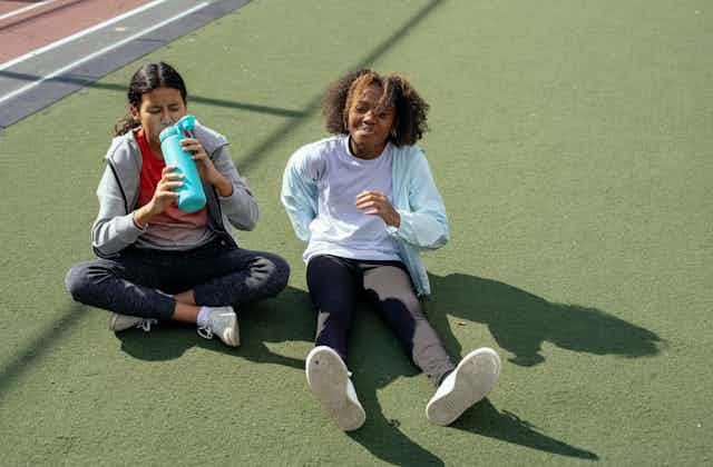 Two teenage girls sit on fake grass in exercise clothing. One is drinking from a water bottle. 