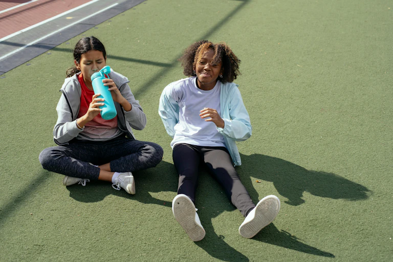 Two teenage girls sit on fake grass in exercise clothing. One is drinking from a water bottle.