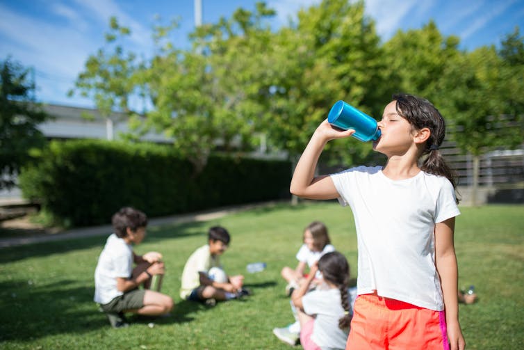 A young girl drinks from a bottle. Children sit on nearby grass.