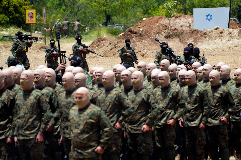 Men in masks and military uniform carrying out a training exercise with armed fighters watching on in the background.