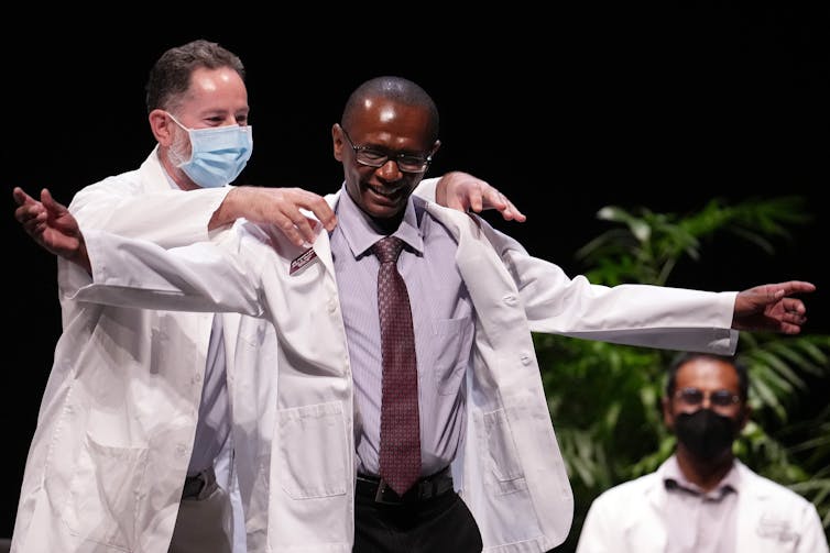A white man wearing a mask is placing a white coat on a Black man who is smiling with his arms outstretched.