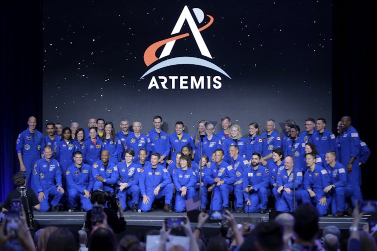 A group of astronauts in blue jumpsuits stand or kneel on a stage in front of a screen displaying the Artemis logo.