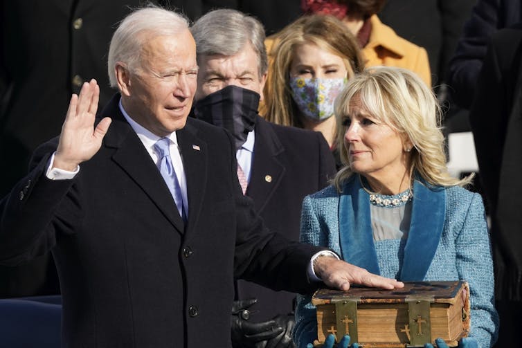 A man with one hand on a large bible, the other raised, speaking.
