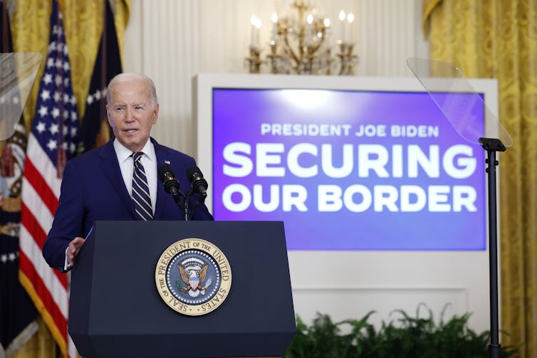 A white man with white hair and a dark blue suit stands at a podium with the presidential seal on it, in front of an American flag and a blue screen that says 'Securing our border'