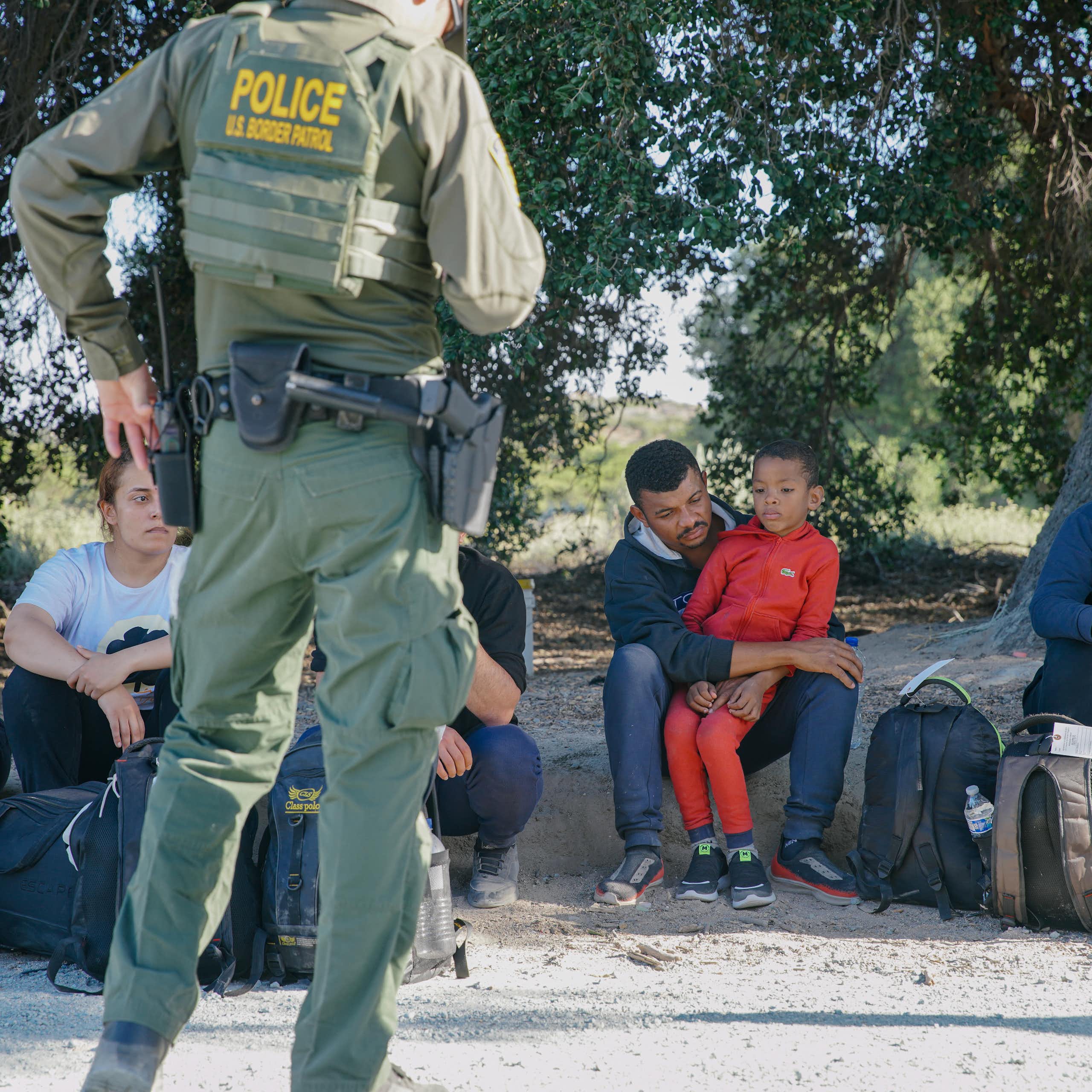 A row of people sit next to backpacks on the ground, as two people wearing green uniforms with the word 'police' on it in yellow stand in front of them.