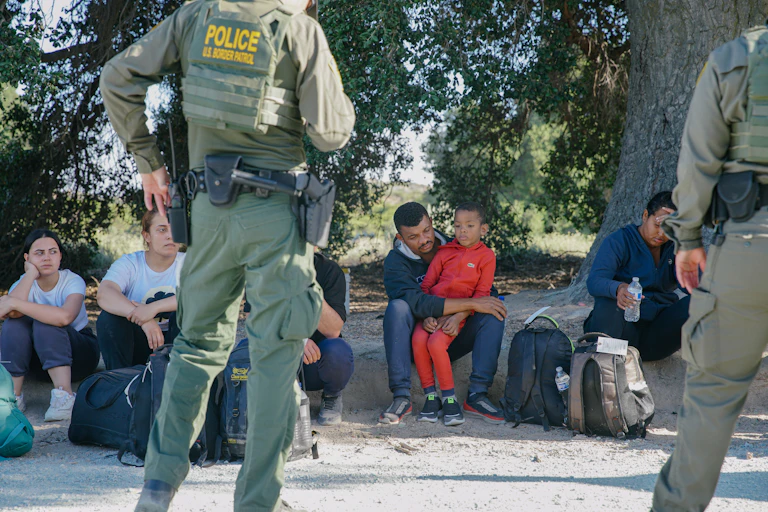 A row of people sit next to backpacks on the ground, as two people wearing green uniforms with the word 'police' on it in yellow stand in front of them.