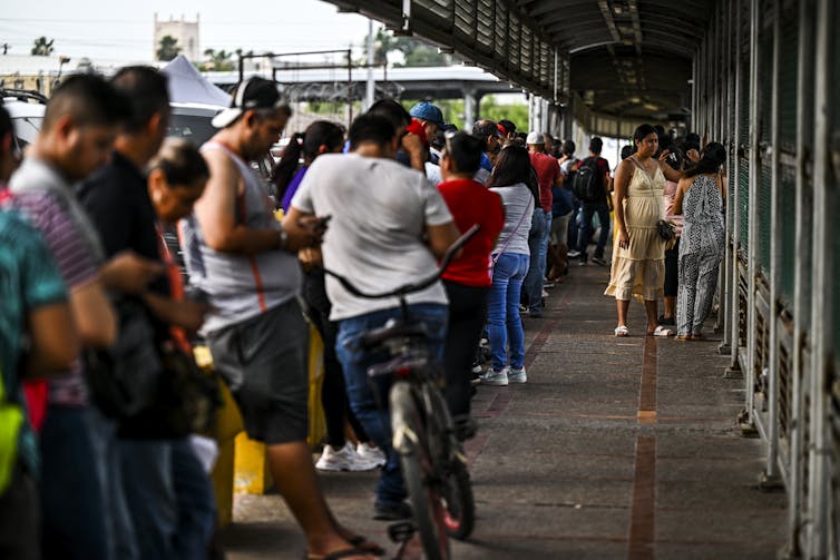 A long line of people stand on the pavement, with a wall next to them.