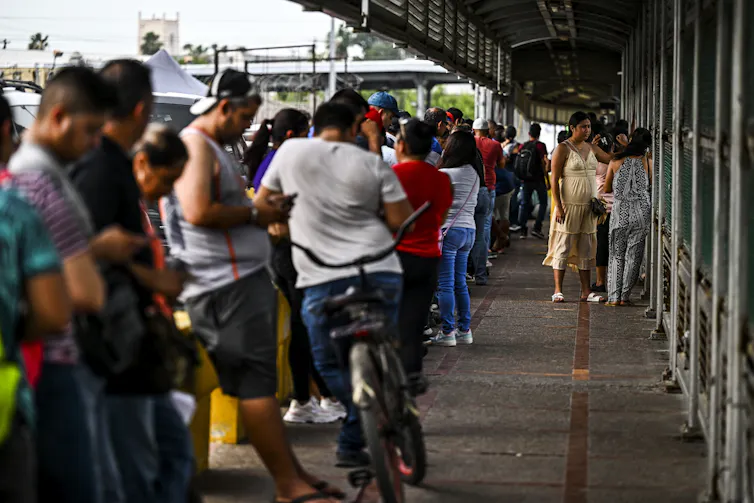 A long line of people stand on the pavement, with a wall next to them.