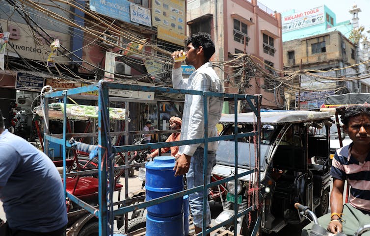 A person stands on a rickshaw drinking water surrounded by traffic and crowds.