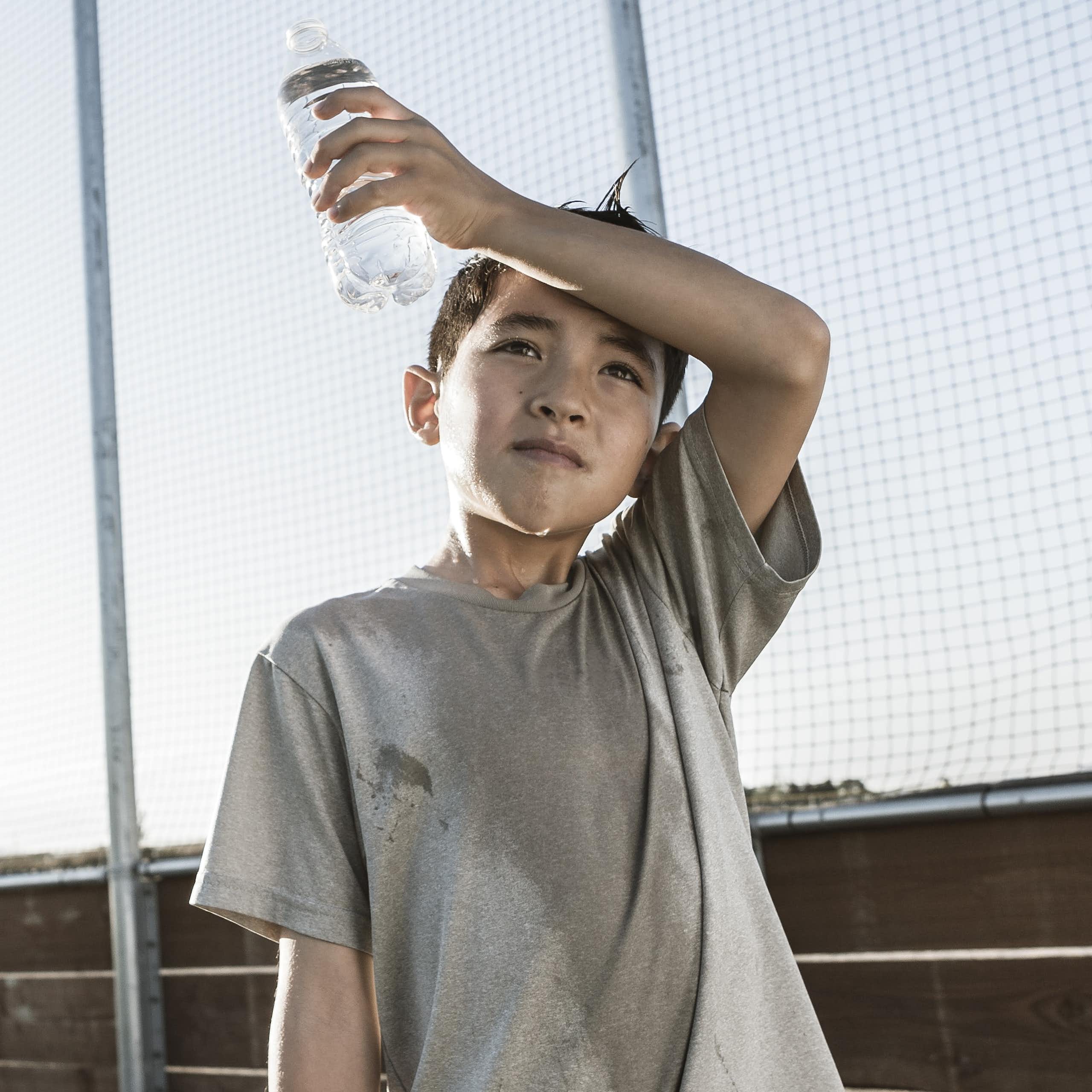 A boy with sweaty shirt wipes sweat off his face while holding a water bottle. A ball field is behind him.