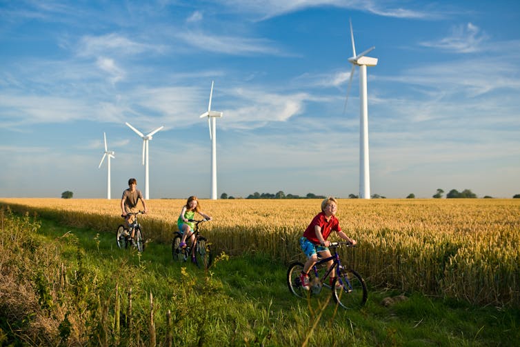 Three children on bicycles ride past a row of wind turbines in a wheat field.