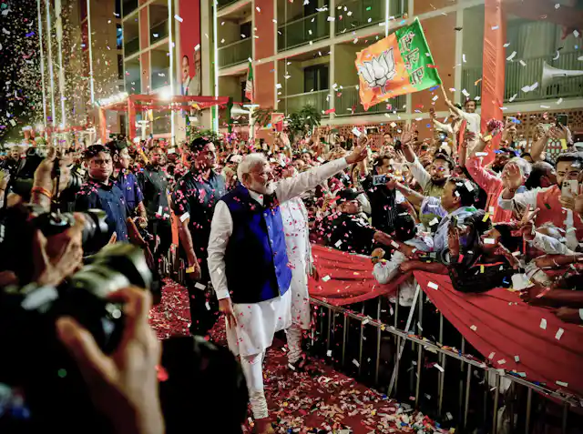 A man in a long white shirt and blue jacket waves to crowds, standing behind a barrier and throwing confetti.