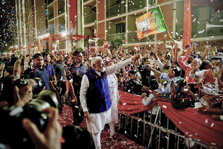 A man in a long white shirt and blue jacket waves to crowds, standing behind a barrier and throwing confetti.