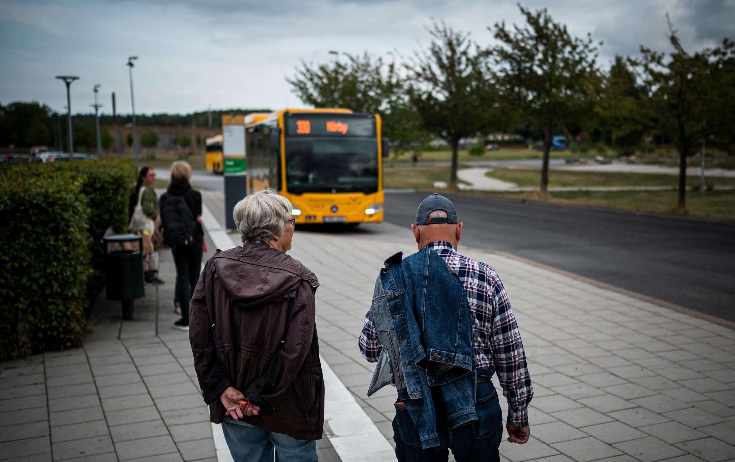 Two older people stand on the street as a bus approaches.