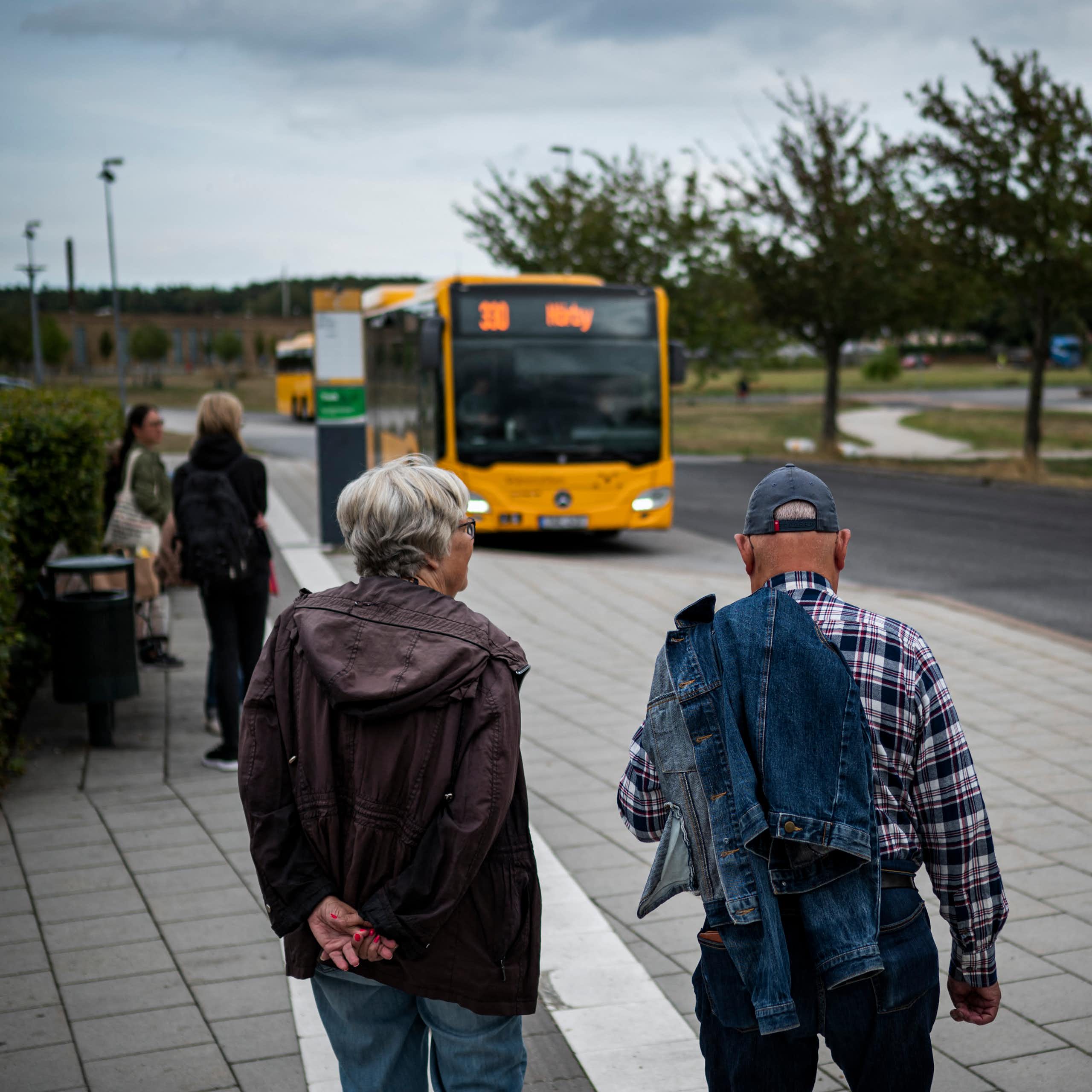 Two older people stand on the street as a bus approaches.
