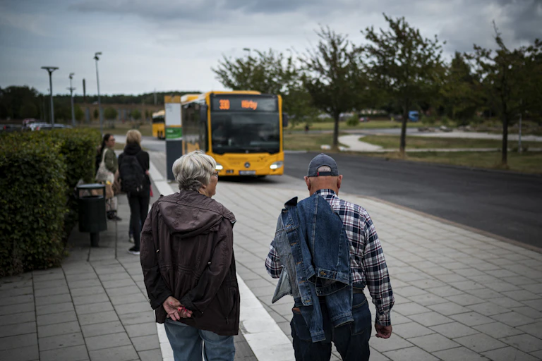 Two older people stand on the street as a bus approaches.