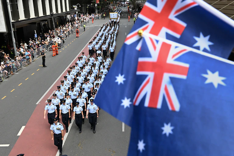 Rows of soldiers march down the street below two Australian flags