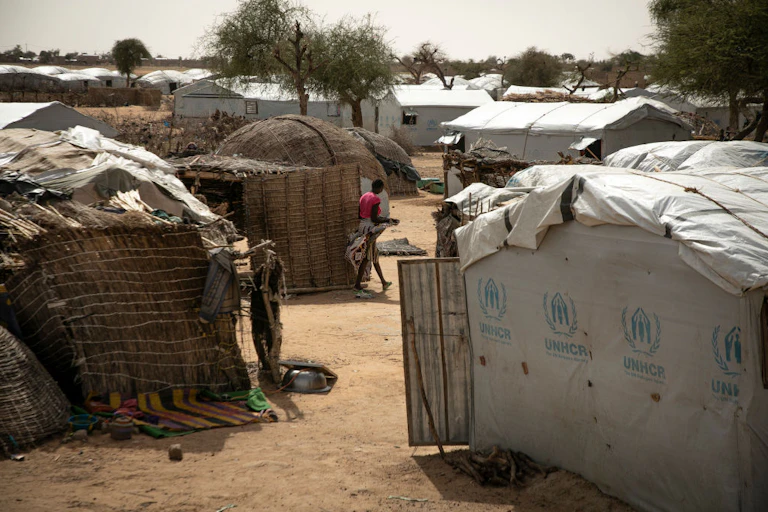 A woman walking between makeshift shelters and tents made with reeds and canvas.
