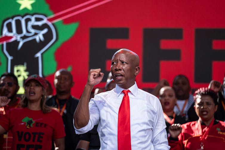 A man wearing a shirt and tie raises a clenched fist with his back to a large banner with the words 'EFF'.
