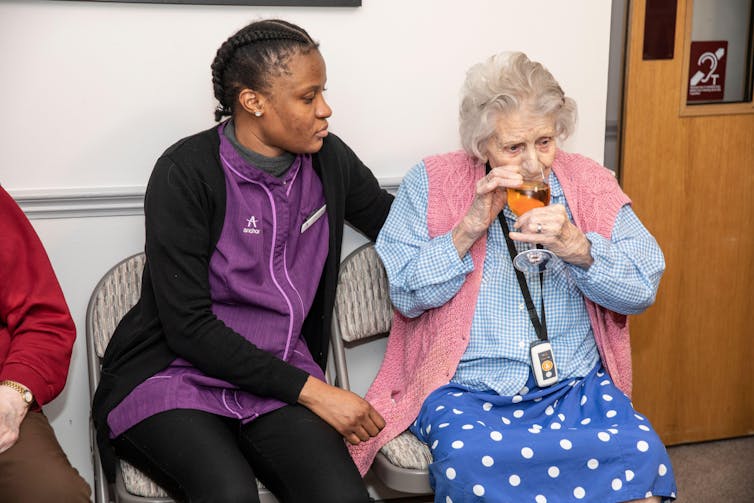 A care worker in a purple uniform sits with an elderly patient in a care home.
