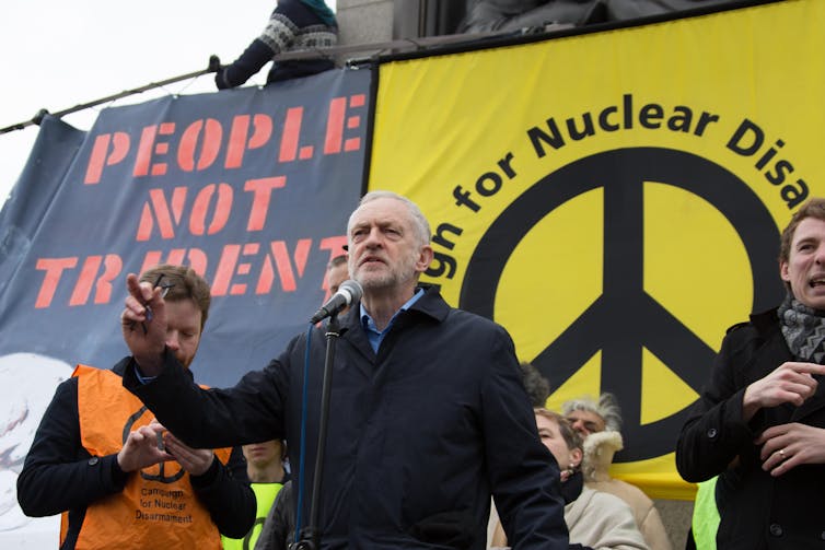 Jeremy Corbyn speaking on stage, with two banners behind him, one reading 'people not trident' and the other with a large peace sign logo for the campaign for nuclear disarmament.
