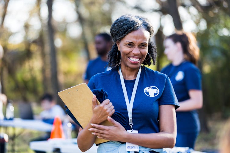 Woman holding clipboard with a badge hanging from a lanyard around her neck smiles during an event where people wearing matching t-shirts.