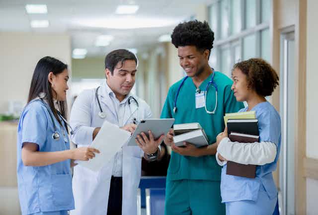 A man in a white coat and stethoscope talking to three young people in scrubs