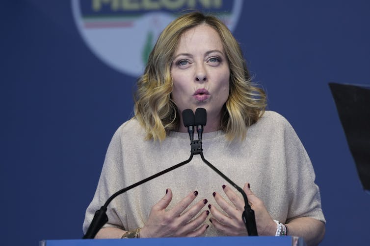 A blonde woman in a beige top speaks into a microphone.