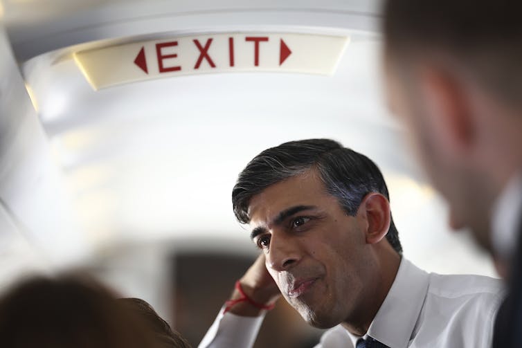 A man sits in an airplane with an exit sign over his head.