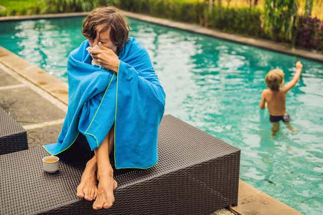 Person wrapped in blanket by poolside blowing nose into tissue