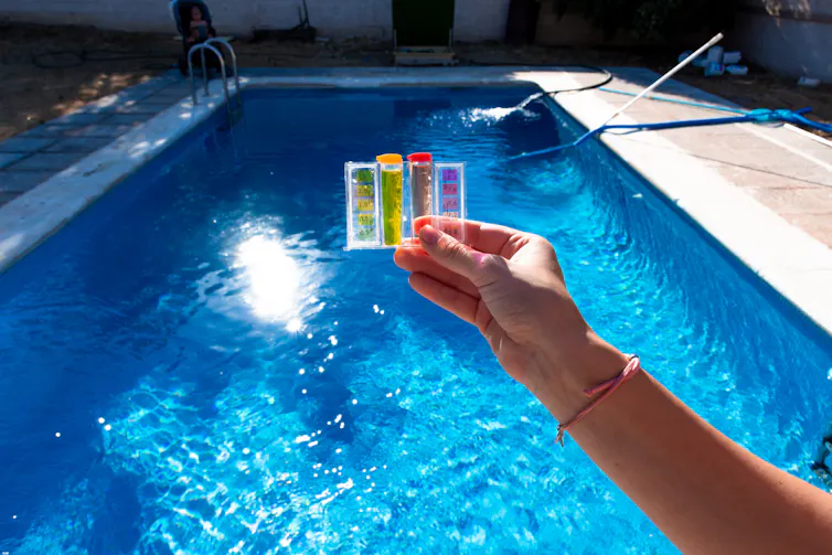 Hand holding pH and chlorine test kit above a swimming pool
