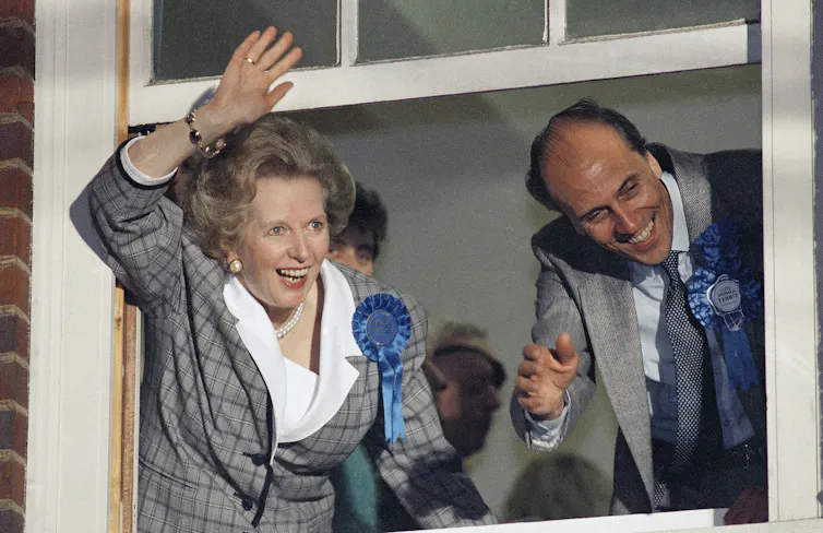 A woman with a bouffant dark blonde hair style smiles broadly and waves out a window.