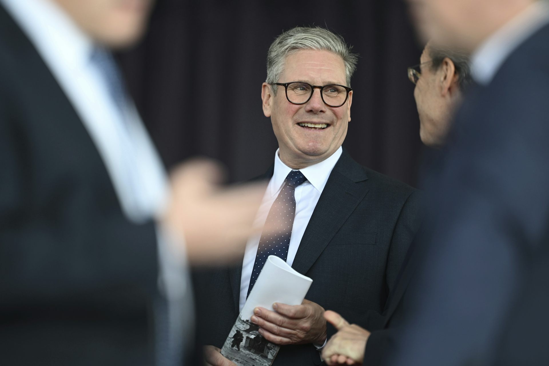 A man wearing glasses and in a dark suit and tie smiles with two blurred men in the foreground standing in front of him.