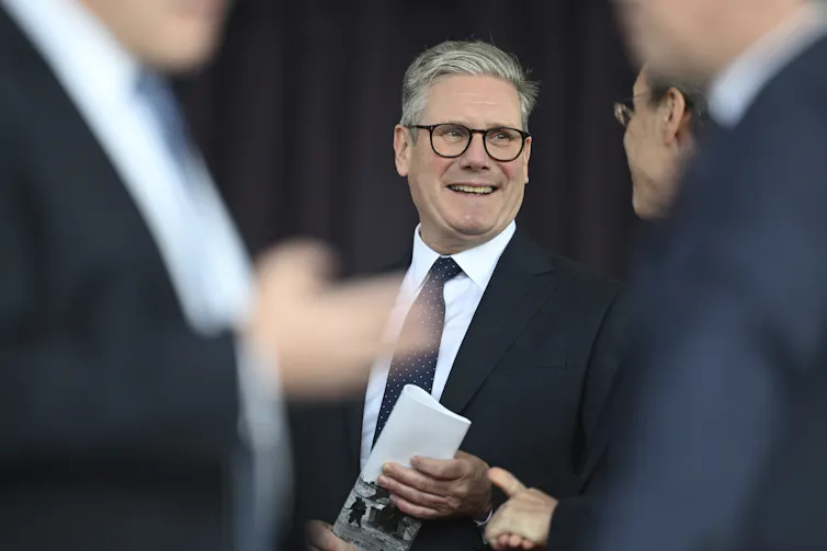 A man wearing glasses and in a dark suit and tie smiles with two blurred men in the foreground standing in front of him.