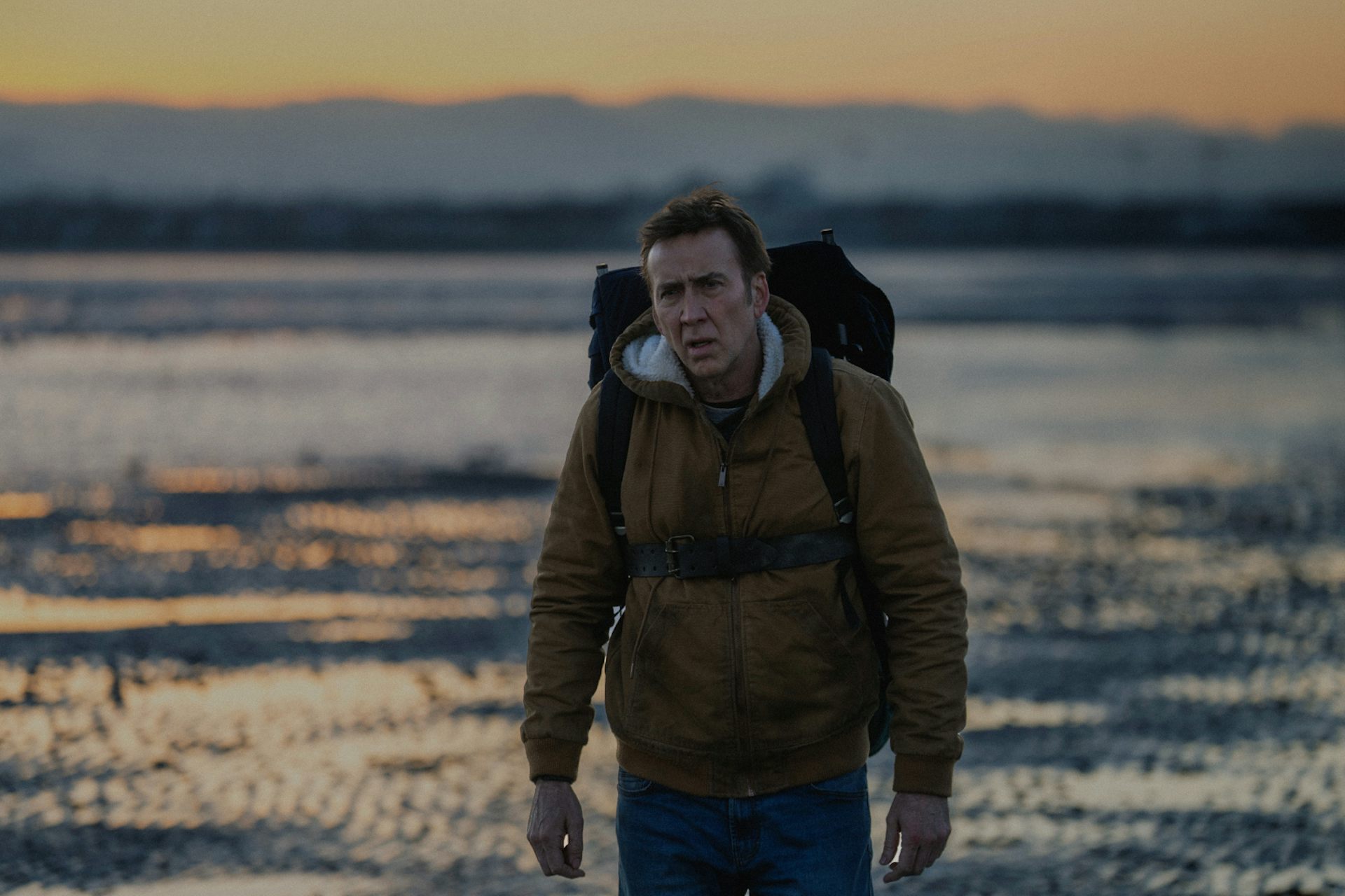 Nicolas Cage wearing a backpack in front of a coastal backdrop.