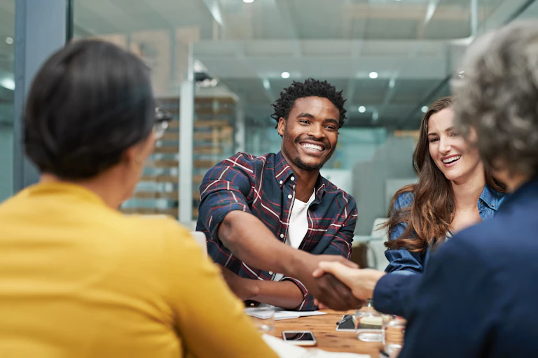 A young man reaching across a table to shake the hand of a person with their back to the camera