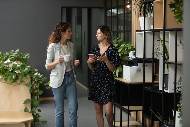 Two young women walking and talking in an office space. One holds a white cup, presumably of coffee or tea.